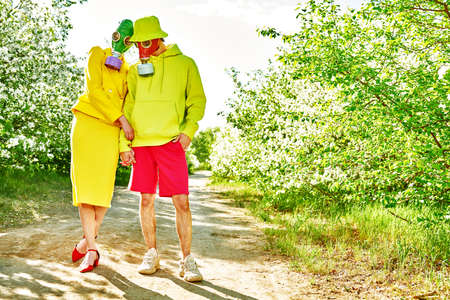A loving man and a woman in bright clothes are walking in the park in gas masks on a sunny day. Air pollution, environmental disaster.の写真素材