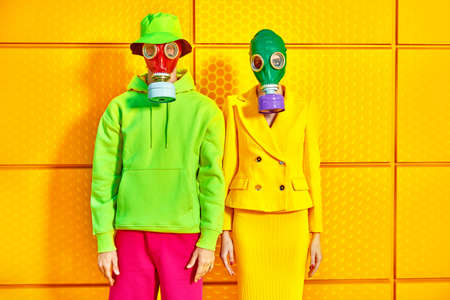 Fashion of the future. Portrait of a woman and a man in bright clothes and gas masks posing by a yellow industrial wall. Environmental disaster, apocalypse.の写真素材