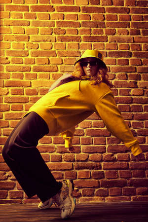 Cool street dancer girl in bright trendy clothes and sunglasses dancing by a brick wall background. Hip-hop dancer.の写真素材