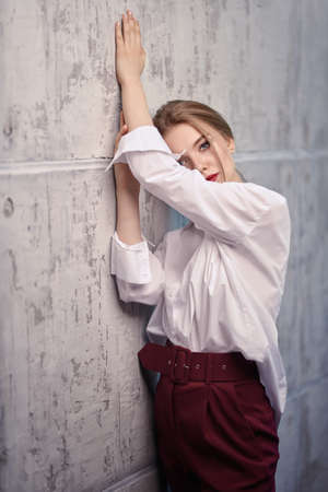 Beauty, fashion portrait. Portrait of a beautiful blonde woman in white blouse and red pants posing at studio against a concrete wall.の写真素材