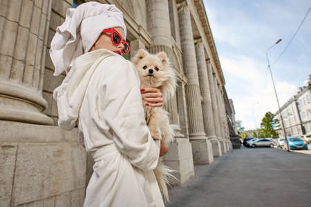 Gorgeous lady in a white bathrobe and elegant red sunglasses walks along the street of a big city with a small dog in her arms. Beauty, fashion.の写真素材