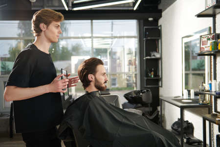 Good looking brunet man getting haircut by hairdresser while sitting in a chair at barbershop.の写真素材