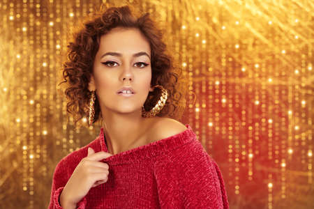 Portrait of a stunning tanned girl with fine curls, bright makeup with arrows and golden earrings. Fashion, cosmetics and jewelry. Golden background with sparkling lights.の写真素材