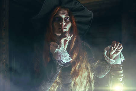 Scary redhead witch with a lantern near the wooden house. Magic, dark force, spell. Halloween.の写真素材