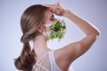 Fashion, beauty and healthcare. Portrait of a beautiful blonde girl posing in a mask decorated with green plants and flowers. Studio portrait.の写真素材