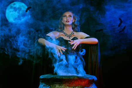 Halloween. A mysterious and gorgeous witch conjures over a cauldron at night on a full moon.の写真素材