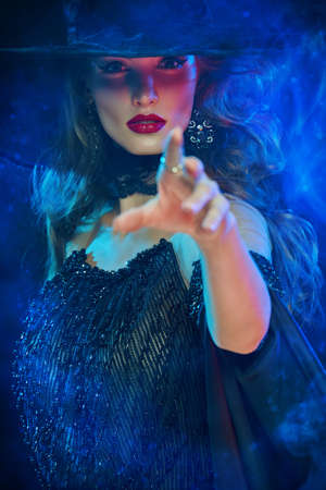 Halloween magic. Gorgeous fairytale witch in black hat and dress bewitches, stretching out her hand to the camera. Halloween party, make-up and costume.の写真素材