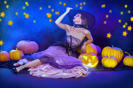 Charming beautiful witch lies with pumpkin lanterns in a luxury evening dress and a hat surrounded by golden stars. Halloween celebration, party. Halloween fashion.の写真素材