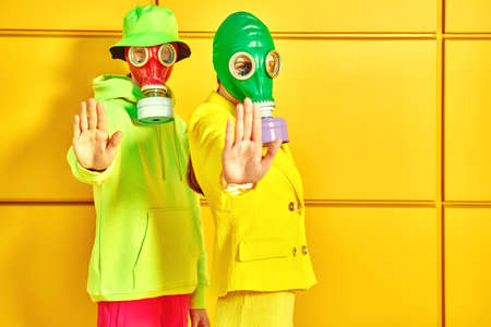 Stop environmental pollution. A couple of modern young people in bright clothes and gas masks show the stop sign by a yellow industrial wall.の写真素材