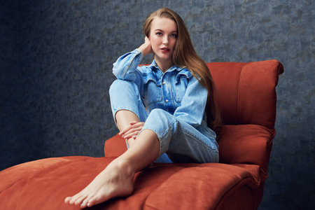 Beautiful girl in denim overalls sits resting on a comfortable couch at home. Modern interior,...