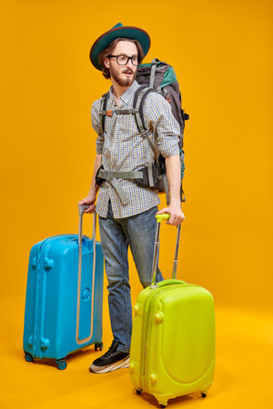 Tourism and travel, vacation. Full length portrait of emotional young hipster man with backpack and suitcases. Studio portrait on a yellow background.の写真素材