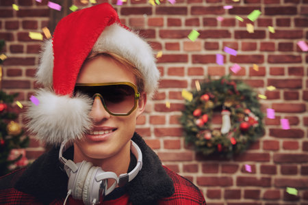 Merry party for Christmas and New Year. Portrait of modern handsome DJ guy in headphones and santa claus hat. Holiday decorations in the background.の写真素材