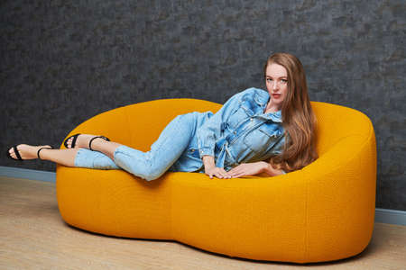 Fashion shot. Beautiful fashion model girl poses in denim overalls on a bright yellow sofa in a modern apartment. Modern interior, furniture.の写真素材