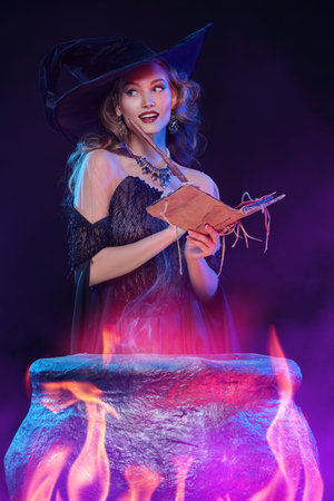Halloween magic. A beautiful young witch in a hat and elegant dress conjures over a cauldron...