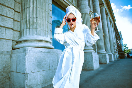 Attractive girl in a white bathrobe and with a white towel on her head walks along a city street...