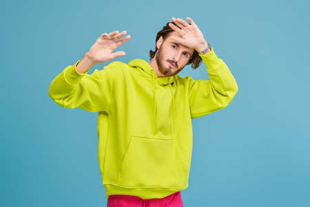 People, emotions. Portrait of a handsome young man in a bright hoodie looking at the camera and covering himself with his hands. Studio portrait on a blue background.の写真素材