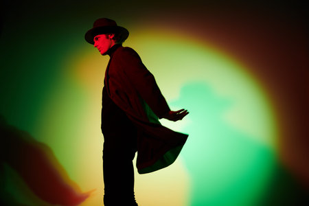 Art portrait. Handsome young man in an elegant hat and a coat posing at studio in mixed color light....