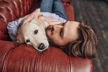 Happy calm young man lies on the couch and hugs his pet Golden Retriever dog.の写真素材