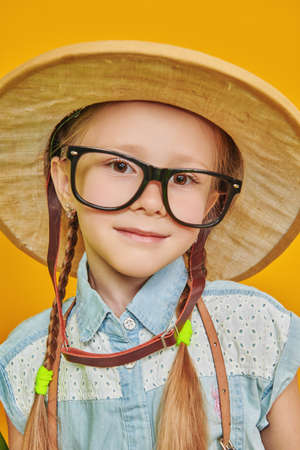 Portrait of a little girl wearing glasses and a traveler's hat smiling at the camera. Children's dreams and fantasies. Studio portrait on a yellow background.の写真素材