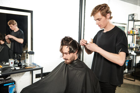 Hairdressing saloon. Handsome brunet man getting haircut by hairdresser while sitting in a chair at barbershop.の写真素材
