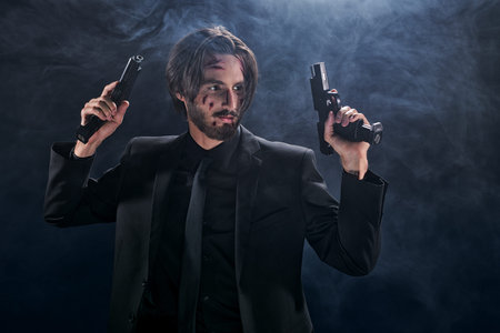 Handsome wounded action hero in a black suit with guns in his hands on a dark foggy background. Copy...