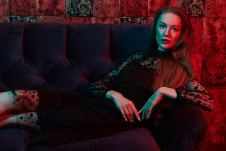 Beautiful woman in black evening dress lies on a velvet sofa in luxury apartments. Glamorous...