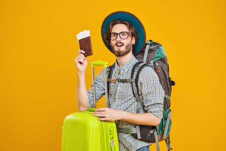 Dreamy young hipster man with backpack and suitcase holding his passport. Tourism and travel, vacation. Studio portrait on a yellow background.の写真素材