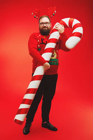 Cheerful fat bearded hipster man wearing deer horns and red sweater posing with huge candy cane and laughs. Red background. Christmas and New Year celebration.の写真素材