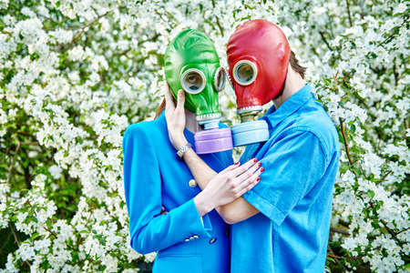 A loving man stands close to his beloved girl in a blossoming apple orchard. They wear blue clothes and bright gas masks. Modern art. Air pollution, environmental disaster.の写真素材