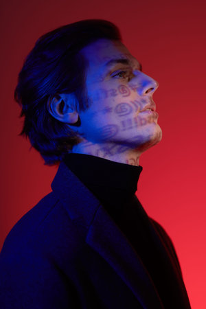 Portrait of a concentrated handsome man in elegant dark clothes posing at studio in dark red and...