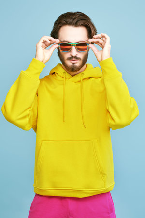 Young man in sunglasses and a bright hoodie poses on studio on a blue background. Youth bright...
