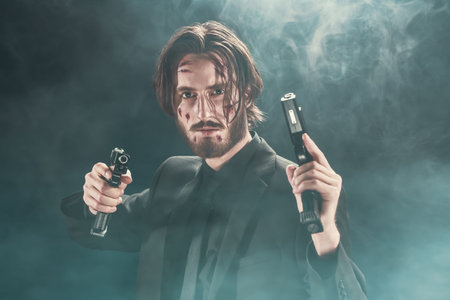 Brave special agent in a black suit with guns in his hands on a dark foggy background.