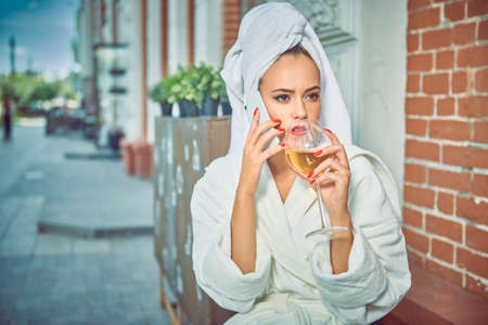 Attractive girl in a white terry dressing gown with a white towel on her head sits at a table in a street cafe with a glass of wine and gossips on the phone. Fashion shot.の写真素材