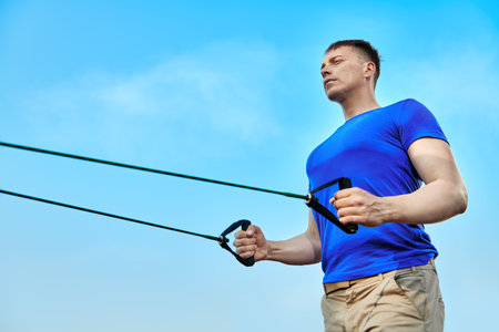 Working out at outdoor gym. An athletic man does strength exercises with elastic bands against the...