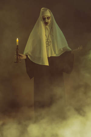 Full length portrait of a scary devilish possessed nun with a candle in her hand standing in a dark room in smoke. Horrors and Halloween.の写真素材