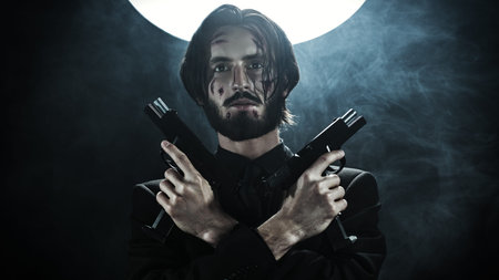 Handsome wounded action hero in a black suit with automatic guns in his hands on a dark foggy...