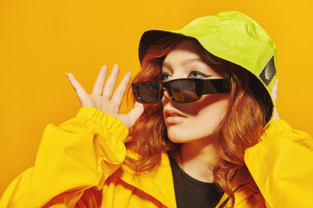 Stylish teenage girl in yellow hoodie and sunglasses looks away on a yellow background at studio....
