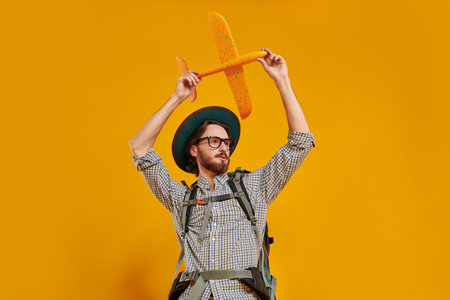 Portrait of a young hipster man in a hat and glasses standing with his backpack and launching a plane into the sky. Tourism and travel, vacation. Studio portrait on a yellow background with copy space.の写真素材