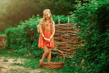 A cute girl dressed in hippie style stands with a basket in the countryside and smiles. Summertime. Kid's fashion.の写真素材