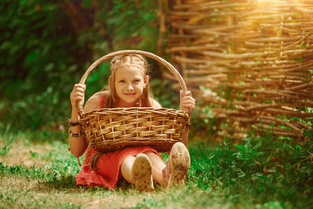 A cheerful little girl sits on the grass with a basket and laughs. Happy summertime.の写真素材