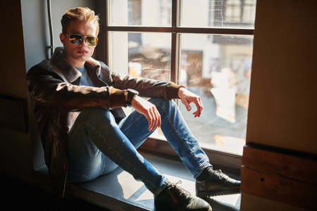 Fashionable young man in black leather jacket sits on the windowsill in apartments. Men's fashion....