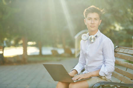 Happy teenage boy sits in a park on a sunny summer day with a laptop and headphones and smiles.の写真素材