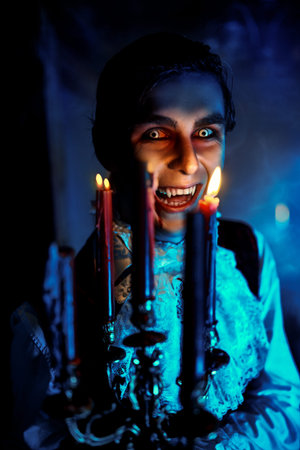 A bloodthirsty vampire aristocrat looks and grins through the candles. 19th century style. Halloween portrait of man.の写真素材