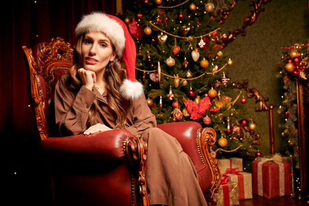 Portrait of an elegant woman in Santa hat sitting in a vintage armchair in a rich Christmas...