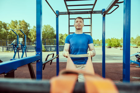 Working out at outdoor gym. Handsome muscular man does strength exercises with elastic band at...