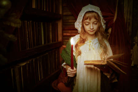 Christmas magic. Beautiful little girl in a nightgown reads a book holding a candle in the home library. Beautiful fairy christmas decoration.の写真素材