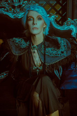 A gorgeous white-haired lady in a rich black dress is posing on the armchair in castle. Portrait in...