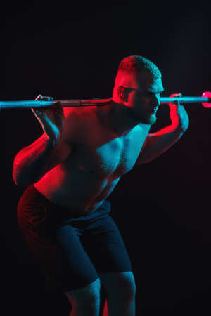 Muscular brutal man doing exercises with barbell in a gym on black background n multicolor red and blue lighting. Health and sports concept.の写真素材