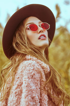 Dreamy young woman in trendy clothes, hat in sunglasses laughs posing next to the reeds on the background of the autumn park. Autumn fashion. Happy people.の写真素材