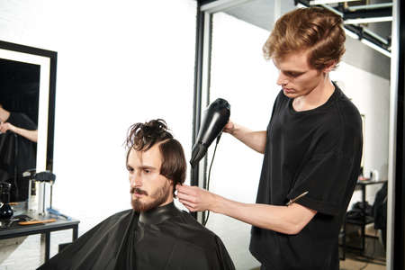 Master barber does the hairstyle and styling with dryer, dries hair to a handsome male client. Barbershop.の写真素材
